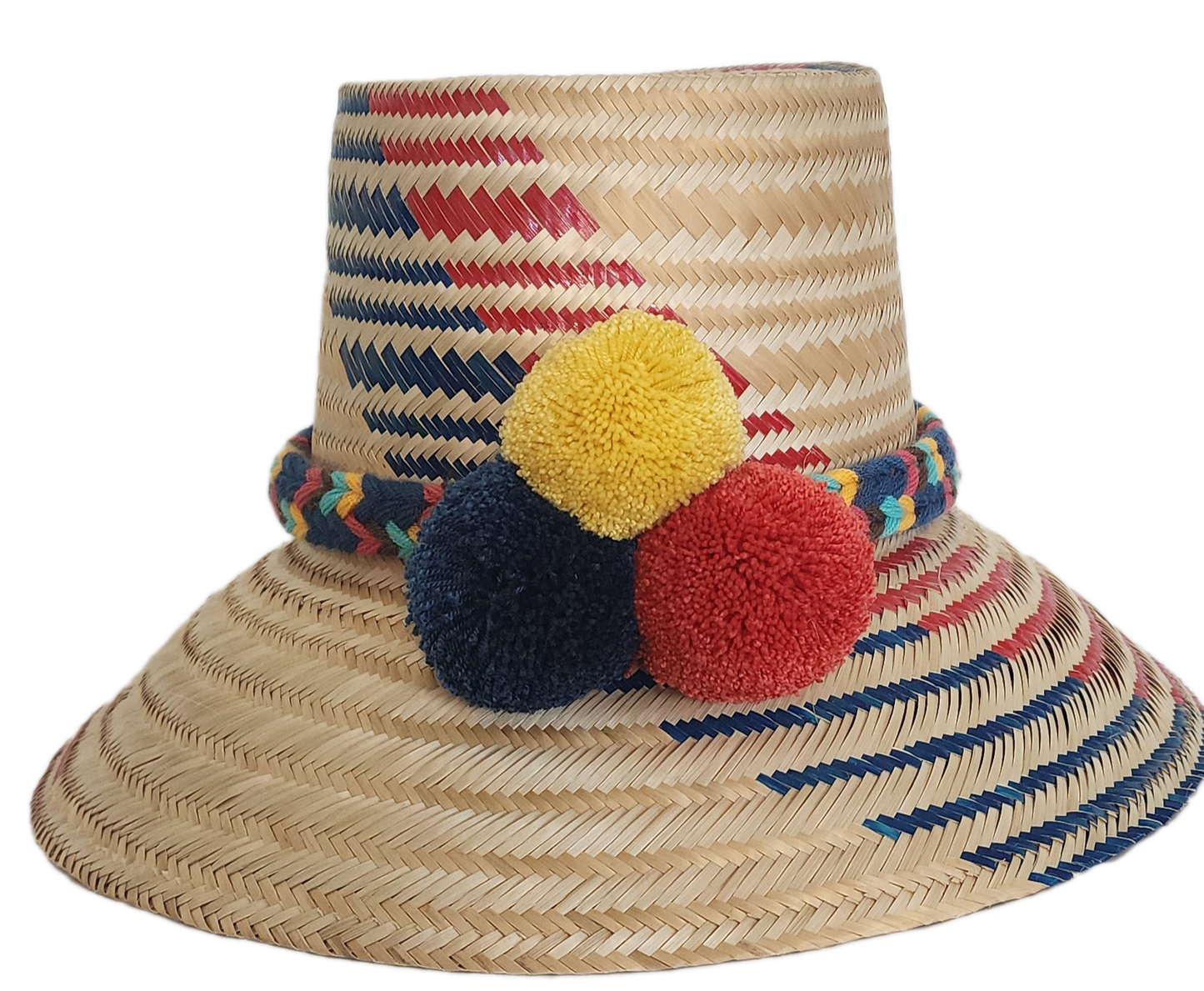 A vibrant, handmade straw hat featuring red and blue geometric patterns on a natural straw base and a colorful pompom band with yellow, dark blue, and red pompoms.
