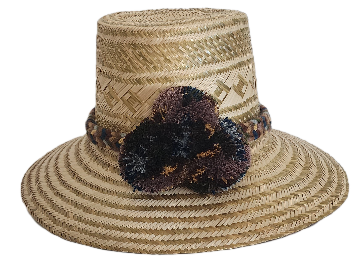 A stylish, handmade straw hat featuring subtle geometric patterns in natural and darker straw, accented by a textured band with brown, blue, and cream elements and a dark, multicolored pompom.