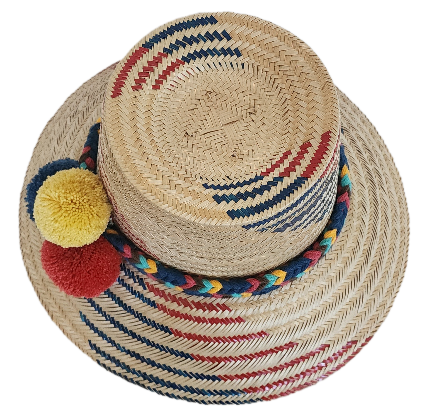A vibrant, handmade straw hat featuring red and blue geometric patterns on a natural straw base and a colorful pompom band with yellow, dark blue, and red pompoms.