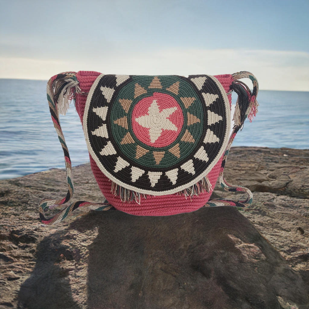 Handmade Wayuu crochet flap bag with starburst pattern in pink, green, beige, and black — colorful boho-chic design with braided strap and fringe.