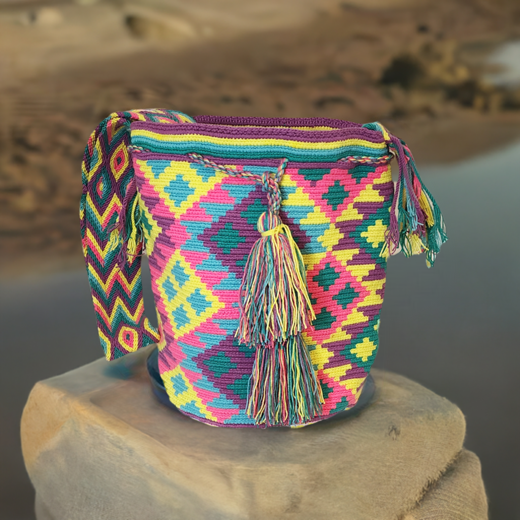 Handmade Wayuu crochet bag with vibrant geometric pattern in pink, teal, yellow, green, and purple tones. Features braided tassels and long strap.