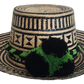 Handcrafted Wayuu straw hat with bold black geometric patterns, natural beige base, a wide circular brim, and a black and green pom-pom band accent.