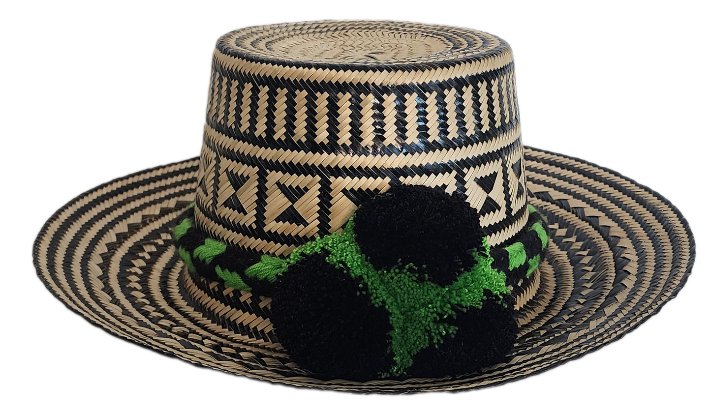 Handcrafted Wayuu straw hat with bold black geometric patterns, natural beige base, a wide circular brim, and a black and green pom-pom band accent.
