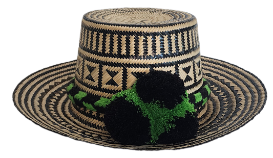 Handcrafted Wayuu straw hat with bold black geometric patterns, natural beige base, a wide circular brim, and a black and green pom-pom band accent.