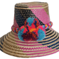 Boho Wayuu straw hat with vibrant pink, navy, and natural woven design, a multicolor braided band, and playful pom-poms in turquoise, red, and orange — a colorful, handcrafted bohemian summer accessory.