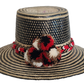 Colorful boho straw hat handwoven by Wayuu artisans, featuring a black and natural pattern, red-and-white braided band, and vibrant pom-poms — a bohemian fashion accessory perfect for summer.