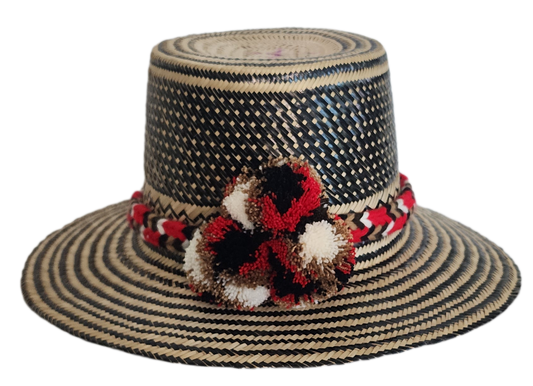 Colorful boho straw hat handwoven by Wayuu artisans, featuring a black and natural pattern, red-and-white braided band, and vibrant pom-poms — a bohemian fashion accessory perfect for summer.