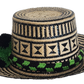 Handcrafted Wayuu straw hat with bold black geometric patterns, natural beige base, a wide circular brim, and a black and green pom-pom band accent.