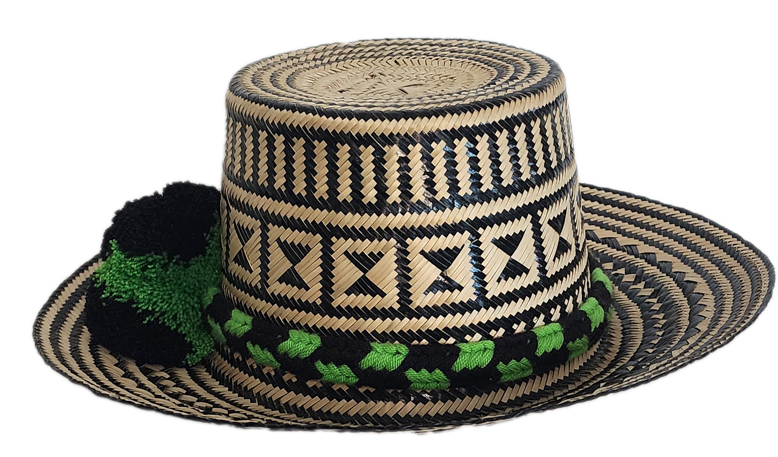 Handcrafted Wayuu straw hat with bold black geometric patterns, natural beige base, a wide circular brim, and a black and green pom-pom band accent.