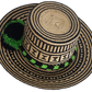 Handcrafted Wayuu straw hat with bold black geometric patterns, natural beige base, a wide circular brim, and a black and green pom-pom band accent.