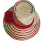 A vibrant, handmade straw hat featuring red and green geometric patterns and a bold red pompom band with three fluffy pompoms.