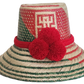 A vibrant, bohemian  handmade straw hat featuring red and green geometric patterns and a bold red pompom band with three fluffy pompoms.