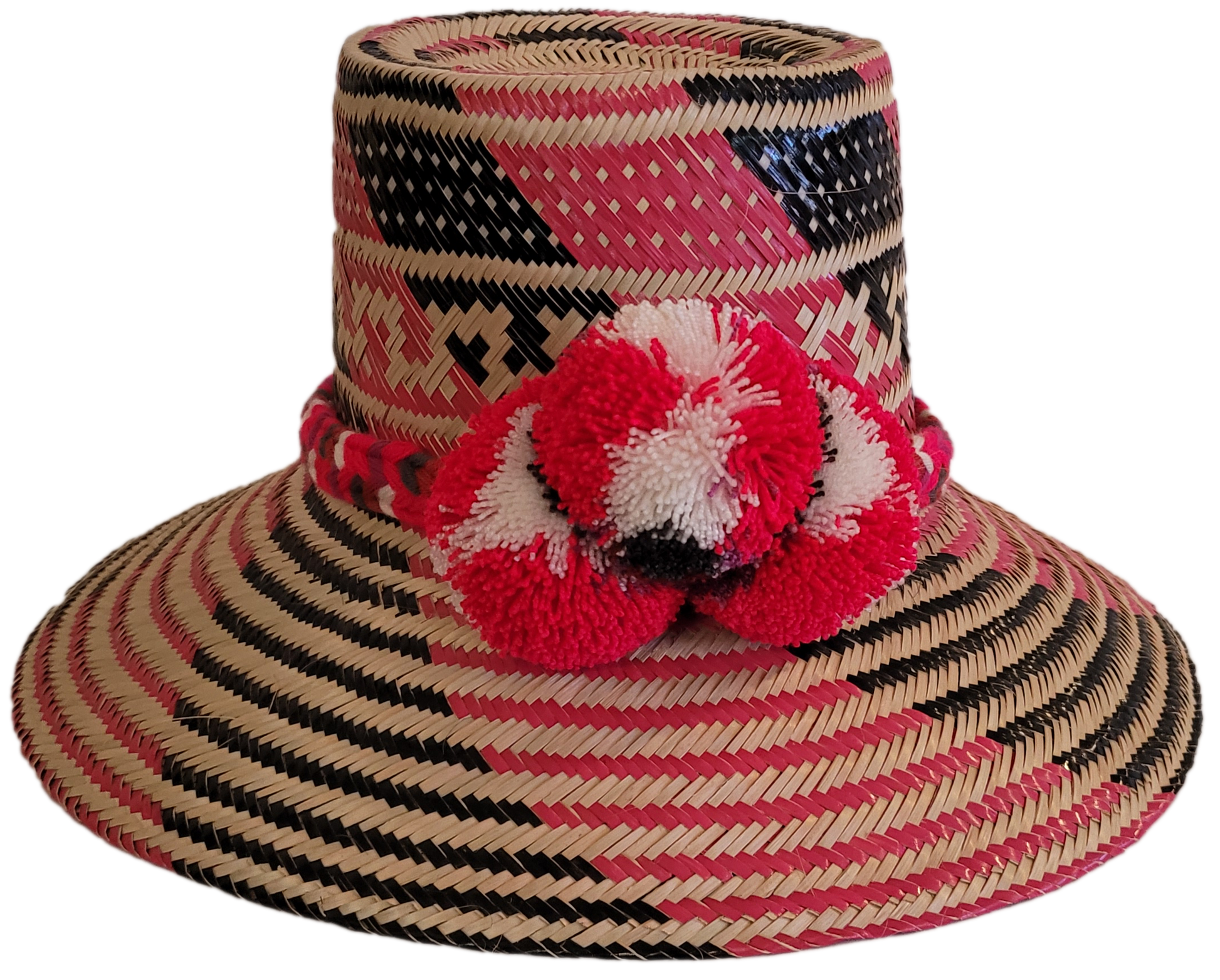 Handmade Wayuu straw hat in pink, black, and beige with red pompoms.

