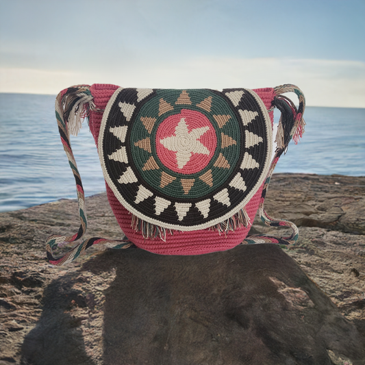 Handmade Wayuu crochet flap bag with starburst pattern in pink, green, beige, and black — colorful boho-chic design with braided strap and fringe.