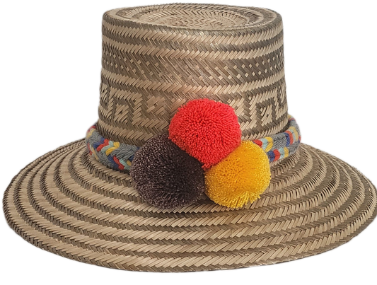 Handwoven Wayuu straw hat with intricate gray-beige tribal patterns, a colorful braided band, and red, yellow, and dark brown pom-poms. Artisan boho style.