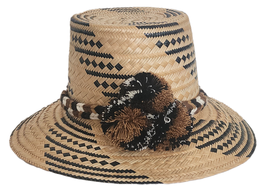Handwoven Wayuu straw hat with black and brown geometric patterns, a braided band, and matching pom-poms. A rich, artisan-crafted boho accessory.