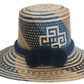 Artisan Wayuu straw hat with bold blue and cream geometric weaving, wide brim, and navy pom-pom band. A handcrafted boho accessory for standout summer style.