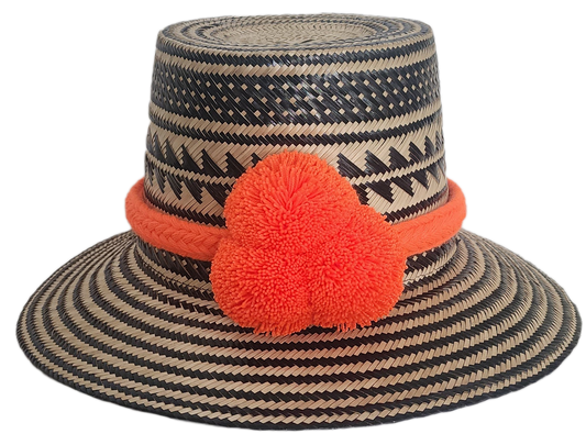 Handwoven Wayuu straw hat with black zigzag tribal patterns and a bold orange pom-pom band. A vibrant artisan boho accessory with modern contrast.