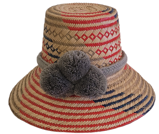 Handwoven Wayuu straw hat with red, navy, and natural patterns, featuring gray pom-poms and a braided band — boho summer accessory.