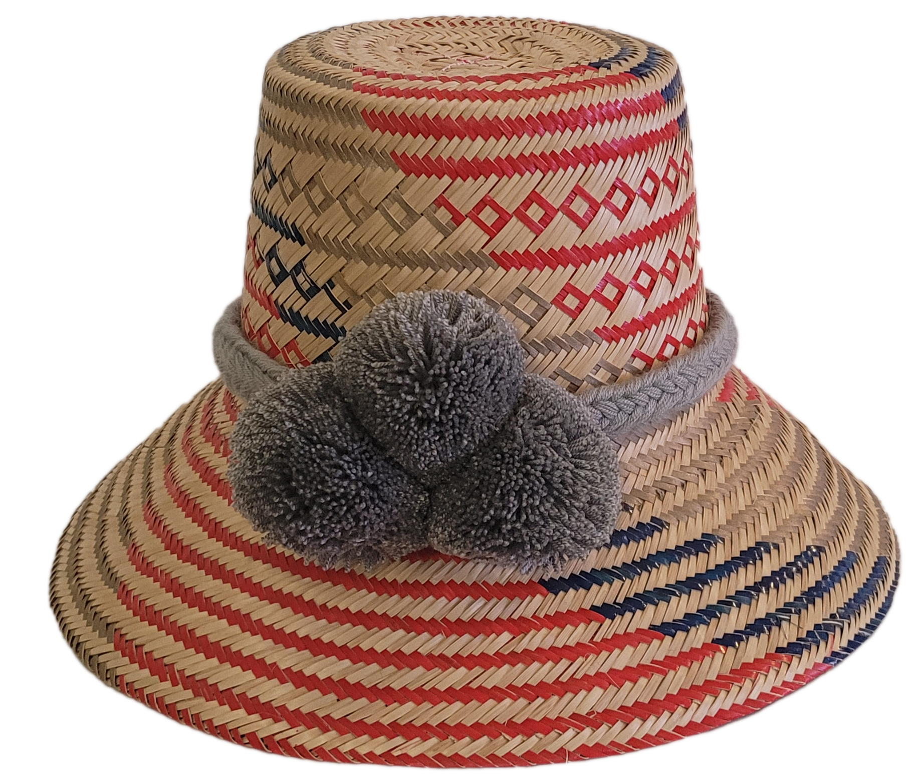 Handwoven Wayuu straw hat with red, navy, and natural patterns, featuring gray pom-poms and a braided band — boho summer accessory.
