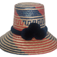Handwoven Wayuu straw hat with bold red and blue geometric patterns, finished with a navy blue braided band and three navy pom-poms in boho colors.