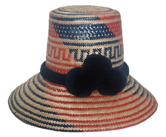 Handwoven Wayuu straw hat with bold red and blue geometric patterns, finished with a navy blue braided band and three navy pom-poms in boho colors.
