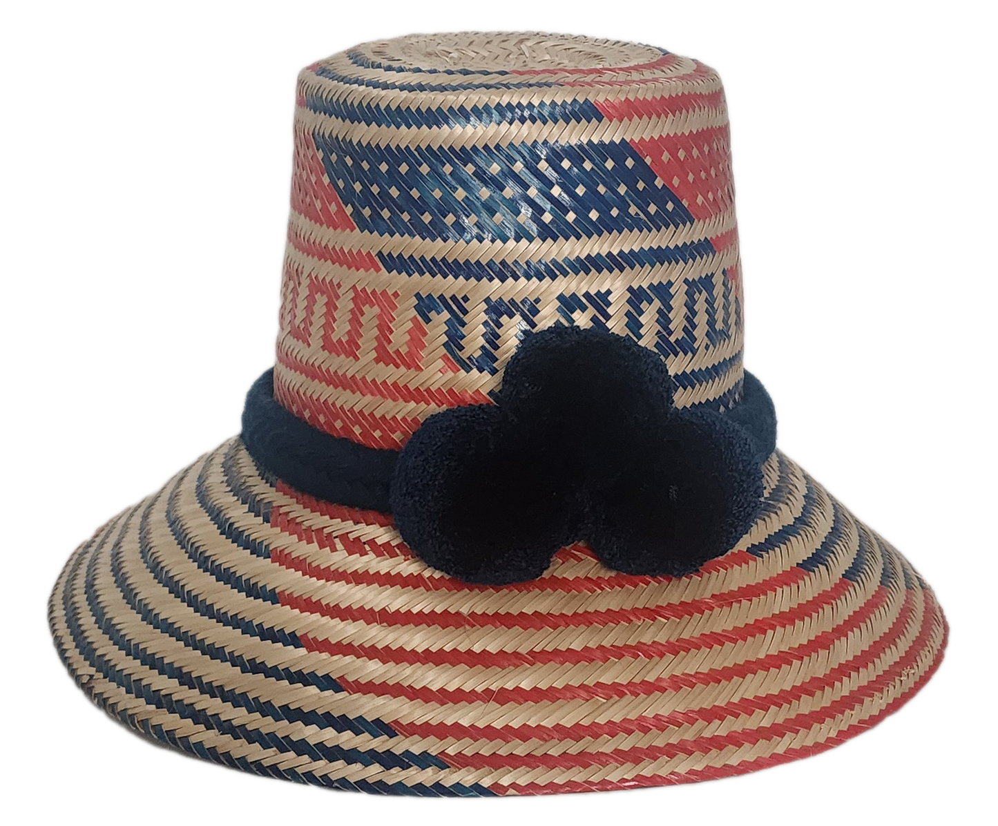 Handwoven Wayuu straw hat with bold red and blue geometric patterns, finished with a navy blue braided band and three navy pom-poms.
