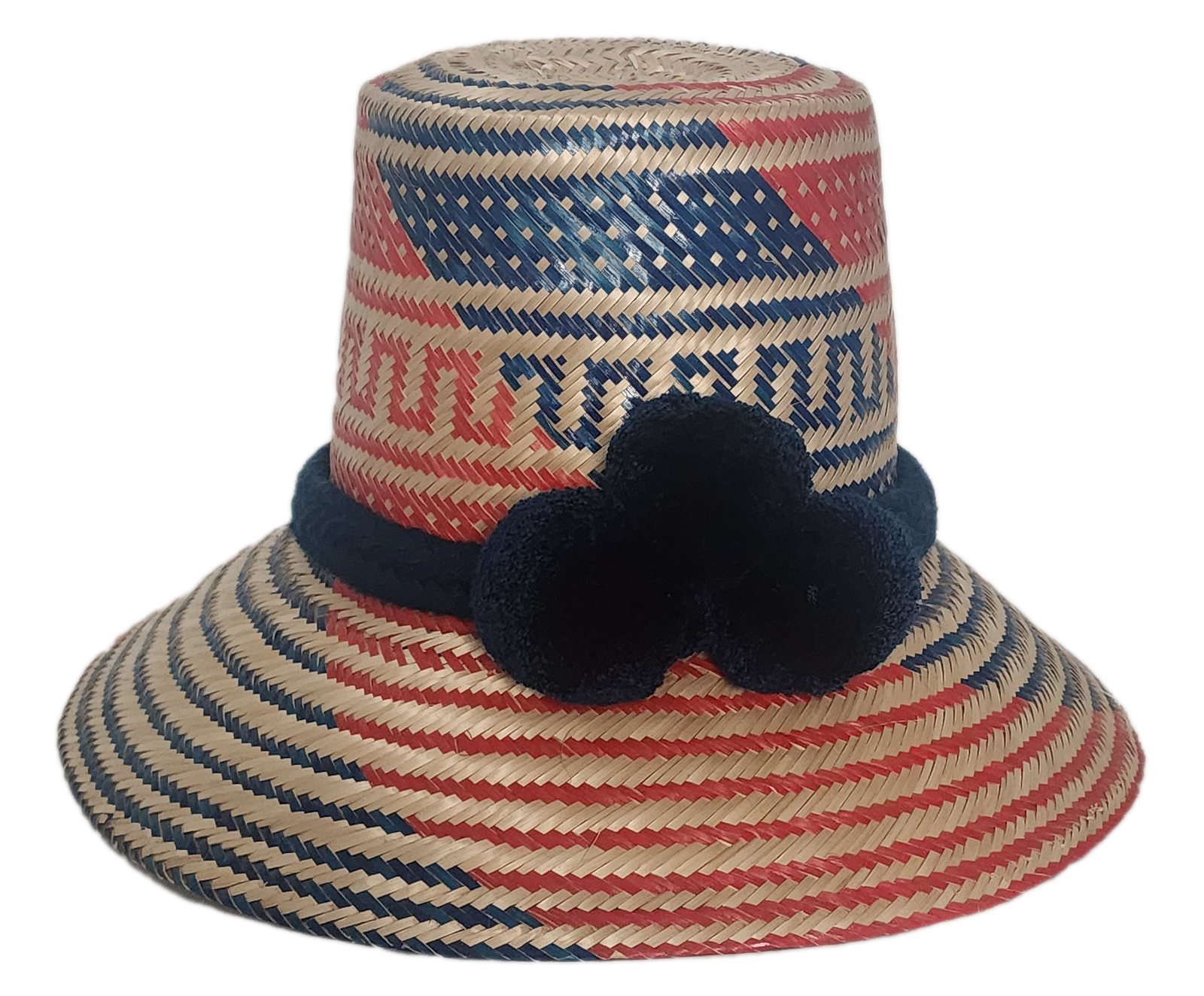 Handwoven Wayuu straw hat with bold red and blue geometric patterns, finished with a navy blue braided band and three navy pom-poms.