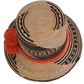 Black and beige Wayuu straw hat with a bright orange braided band and pompoms — a colorful, handmade boho accessory full of artisan charm.