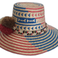 Boho straw hat with bold red and blue patterns, braided band, and colorful pompoms.