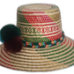 Handmade Wayuu straw hat with red and green woven patterns, colorful braided band, and deep-toned pompoms.